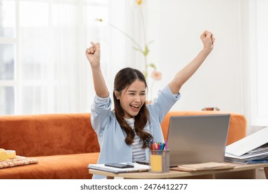 Asian young woman joyfully raises her arms in celebration while working on her laptop at home,moment of professional success, reflecting the positive side of remote work and effective goal accomplish - Powered by Shutterstock