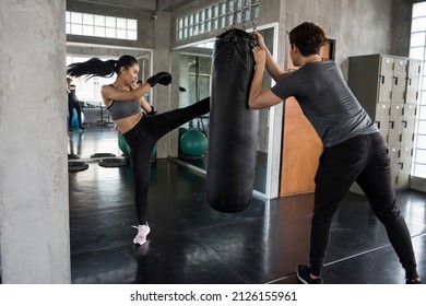 Asian young woman kick boxing sandbag held by personal trainer at sport fitness gym. Athletic boxer practice Muay Thai with coach for bodybuilding and healthy lifestyle concept.