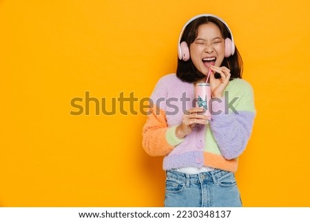 Asian young woman listening music laughing while drinking lemonade isolated over yellow wall