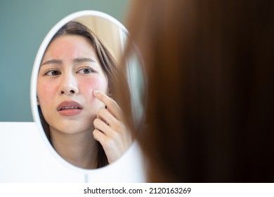 Asian young woman looking face skin in the mirror have a red rash on her face from cosmetic allergy - Shutterstock ID 2120163269
