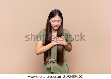 asian young woman looking sad, hurt and heartbroken, holding both hands close to heart, crying and feeling depressed