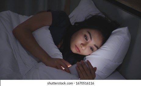 Asian Young Woman Lying On Bed Play Smartphones During Night Time In Bedroom That Turns Off Lights. Read Social On Mobile Phone. Using Phone In Low Light Has Health Effect On Eyes. Insomnia Concept