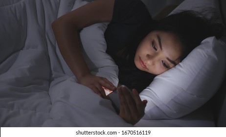 Asian Young Woman Lying On Bed Play Smartphones During Night Time In Bedroom That Turns Off Lights. Read Social On Mobile Phone. Using Phone In Low Light Has Health Effect On Eyes. Insomnia Concept