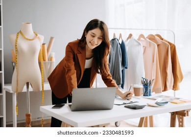  Asian young woman on desk in office of fashion designer and holds tablet, laptop and smartphone on desk - Powered by Shutterstock