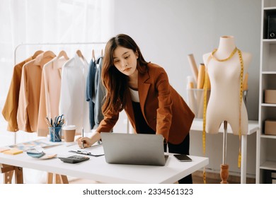  Asian young woman on desk in office of fashion designer and holds tablet, laptop and smartphone on desk - Powered by Shutterstock