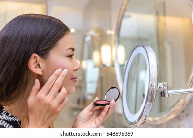 Asian young woman putting blush cream using fingers while looking in makeup mirror at home in bathroom. Morning routine of girl applying make-up before going to work. Face skincare.