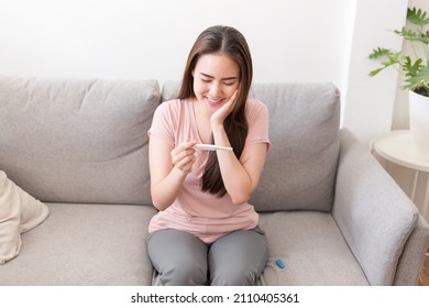 Asian Young Woman Sits On The Sofa Holding Pregnancy Test Smiling See Positive Two Stripes Result. Healthy Female, Ovulation Period, A Modern Device For Detecting Gestation Concept.