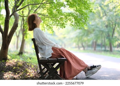 Asian young woman sitting bench look up under green tree in park on sunny day. Full length side view - Powered by Shutterstock