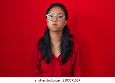 Asian Young Woman Standing Over Red Background Puffing Cheeks With Funny Face. Mouth Inflated With Air, Crazy Expression. 