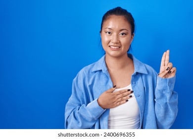 Asian young woman standing over blue background smiling swearing with hand on chest and fingers up, making a loyalty promise oath 