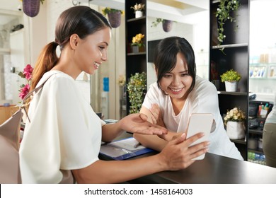 Asian Young Woman Standing At Reception And Showing Photos On Her Mobile Phone To Receptionist In Beauty Salon