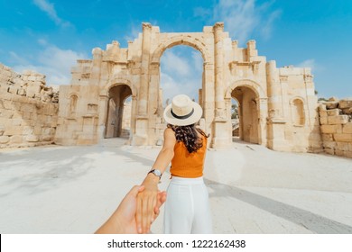 Asian young woman tourist in color dress and hat leading man to South gate of the Ancient Roman city of Gerasa, modern Jerash, Jordan. Traveling together. Follow me.