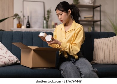 Asian Young Woman Unpacking Cardboard Box Holding Cosmetics Jars Receiving Beauty Products Sitting On Couch At Home. Cosmetology And Selfcare, Delivery Service Concept