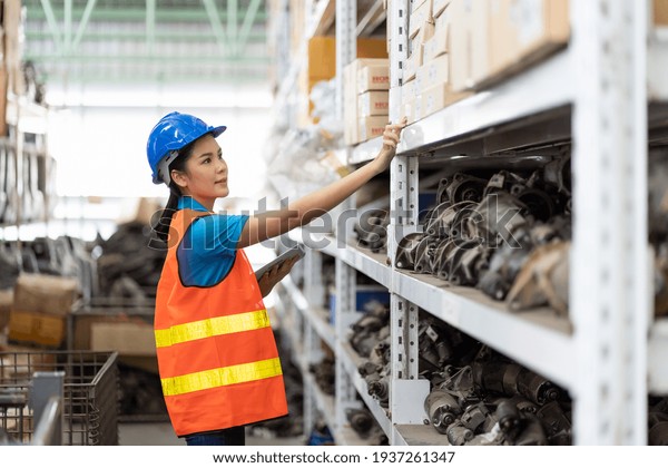 Asian young woman warehouse worker in safety\
vest and helmet using digital tablet for checking barcodes\
automotive spare parts on parcel goods on shelf pallet in\
industrial factory\
warehouse