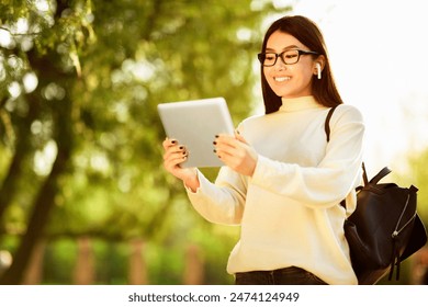 Asian young woman wearing glasses and wireless earbuds uses a tablet while standing in a sunny park. She has a backpack and is smiling, enjoying the outdoor environment. - Powered by Shutterstock