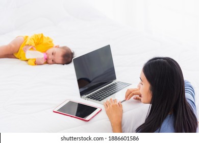 Asian Young Woman Is Working Busy On Laptop Computer And Tablet At Home Office In Bedroom While Her Little Baby Is Sleeping Feeding Lying On The White Bed Lying Near Mom