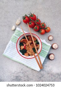 Asian/Chinese/Thai noodles in a bowl with chicken and vegetables. Bowl on a green cloth, mushrooms and  tomatoes. Chopsticks. Traditional meal on a concrete stone background. Top view. Asian dinner