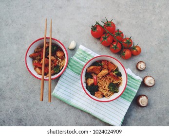 Asian/Chinese/Thai noodles in a bowl with chicken and vegetables. Bowl on a green cloth, mushrooms and  tomatoes. Chopsticks. Traditional meal on a concrete stone background. Top view. Asian dinner