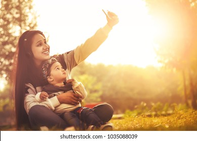 Asian/Indian Beautiful Mother Taking Selfie With Her Smart-phone Camera Of Her And Her Baby Daughter. Outdoor In Nature. 