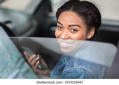 Ask for directions from passerby, travel route and road trip plan. Smiling young african american woman driver holding map and phone, sitting in car, looking through half open window, close up