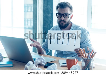 Asking for help. Frustrated young beard man holding piece of paper and asking for help while sitting at his working place in office 