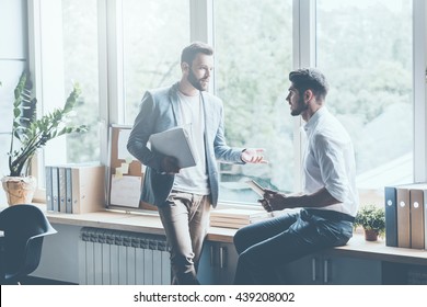 Asking For Professional Advice. Two Young Businessmen In Smart Casual Wear Talking And Gesturing While Leaning At The Window Sill In Office 