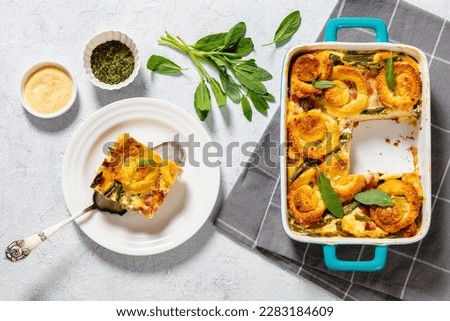 asparagus ham strata topped with sponge cake circles in baking dish on concrete table with ingredients horizontal view from above, flat lay