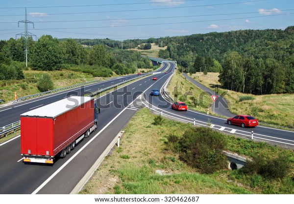 Asphalt highway with red truck and red passenger\
cars in wooded country. Slip road with traffic sign give way.\
Electronic toll gate in the distance. View from above. Sunny summer\
day with blue skies.