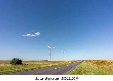 Asphalt Path To The Horizon Surrounded By Electricity Generating Windmills. Blue Sky With A Single Cloud