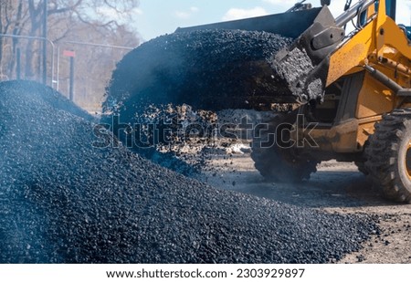 Asphalt paver filled with hot tarmac laying new road surface on new residential housing development site and roadworker operator in orange hi-viz  