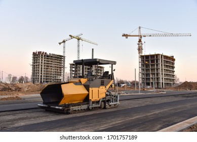 Asphalt Paver Machine During Road Works. Road Machinery At Construction Site. Paving Roller Machine On Paving Works. Tower Cranes Constructing A New Residential Building On Sunset