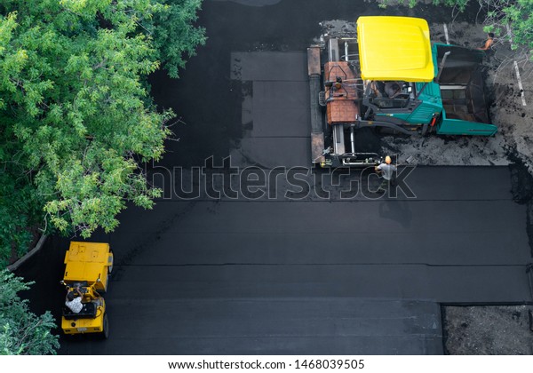 asphalt paver, skating rink and loader are\
working to repair road. Laying new asphalt, top view. Workers\
manage paver and repair road surface in\
yard.