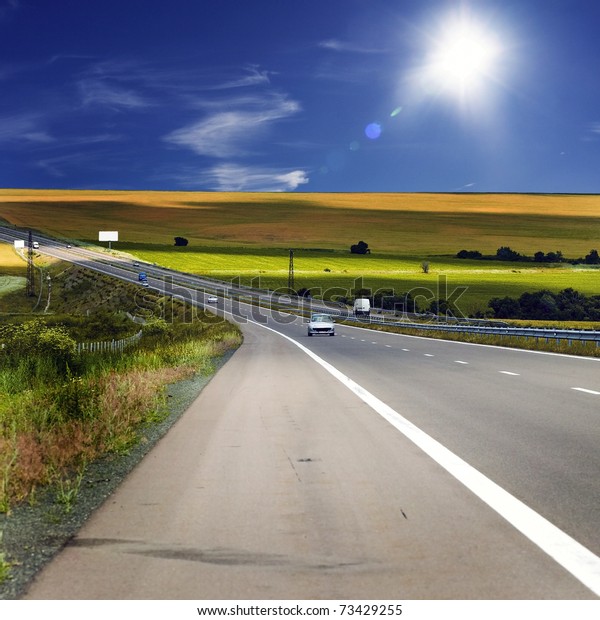 asphalt road with cars against the backdrop of\
arable land and blue sky with\
sun