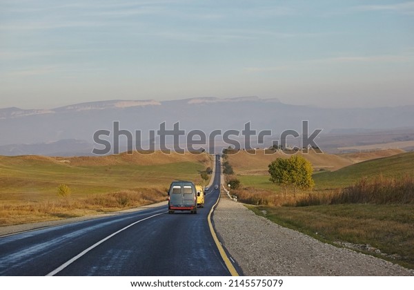 Asphalt road, cars ride on\
the background of a mountain landscape and fields, the sky on a\
sunny day