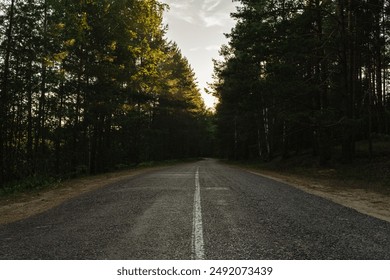 Asphalt road in a dark forest, the tops of the pine trees are illuminated by the setting sun. Slightly grainy retro style photo - Powered by Shutterstock