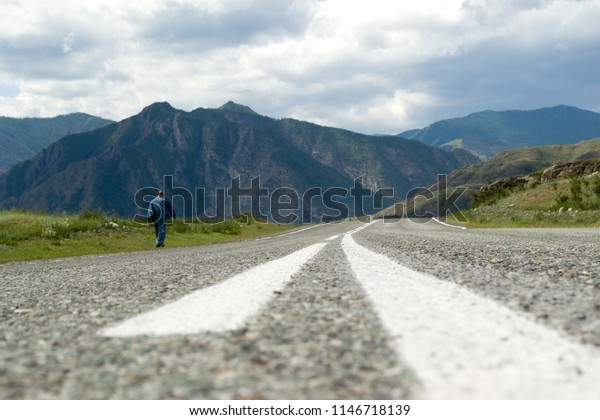 An asphalt road with a dividing\
strip shot close-up and a man, a lone traveler walking along the\
edge of the road. The road leading to the mountains.\
Altai