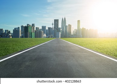 Asphalt Road Leading To A City With Tall Buildings Through Green Meadow