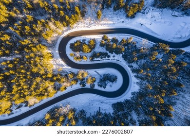 Asphalt road serpentine in snowy wintertime. Cold winter and sunny day above forest road with illuminated trees by rising sun.