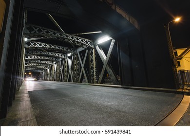 Asphalt Road Under The Steel Construction Of A Bridge In The City. Night Urban Scene With Car Light Trails In The Tunnel. Toned