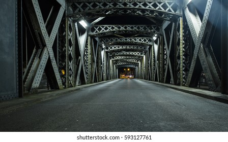 Asphalt Road Under The Steel Construction Of A Bridge In The City. Night Urban Scene With Car Light Trails In The Tunnel. Toned