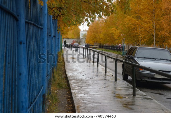An\
asphalted pavement with puddles wet from rain, a bright blue iron\
fence, trees, birches with yellow and green leaves. Silhouette of a\
man, parked car. City courtyard. Autumn\
background
