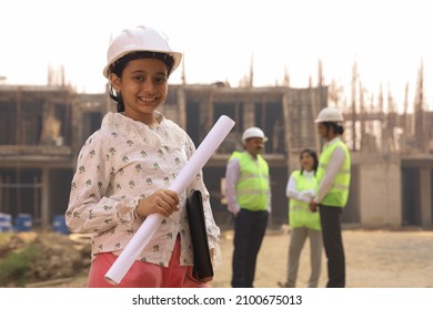 Aspirational happy Indian girl child standing on a construction site holding blue prints and file and wearing engineer's helmet. Portraying to be a future engineer, all the engineers standing behind. - Shutterstock ID 2100675013