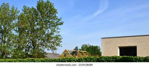 Assembling Kit Tiny Garden House. Neighbor Standing On A Ladder On A Bright Sunny Sunday Morning Building The Roof Of His Temporary Mobile Tiny House Construction Seen From Garden Fence