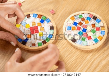 Assembling mosaic coasters at home as a DIY craft project, attaching small moisaic components. Captures focused posture, slow craft rhythm, and mindful artistic attention.