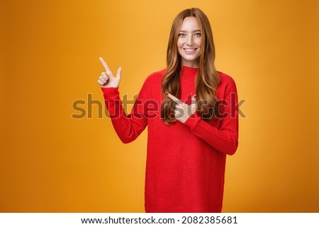 Assertive and helpful charming young female assitant in red knitted dress with ginger hair smiling friendly and pleasant as pointing at upper left corner, showing interesting thing over orange wall