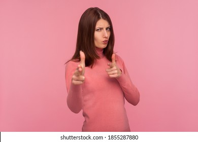 Assertive self confident woman with brown hair in pink sweater showing guns with her fingers seriously looking at camera aiming you, self defence. Indoor studio shot isolated on pink background