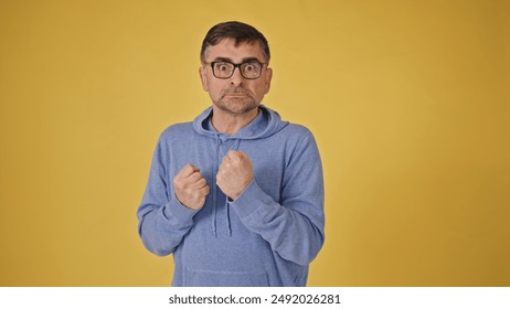 Assertive senior man in glasses making threatening gesture against yellow background