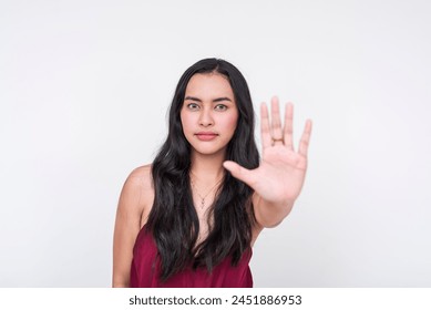 Assertive young Asian lady in chic maroon dress, firmly indicating stop with her hand. Isolated on a clean white background.