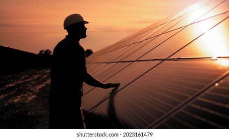 Assistance Technical Worker In Uniform Is Checking An Operation And Efficiency Performance Of Photovoltaic Solar Panels. Unidentified Solar Power Engineer Touches Solar Panels With His Hand At Sunset