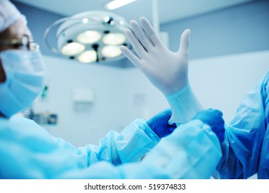 Assistant helps the surgeon put on latex gloves before the operation. A team of surgeons in the operating lamp background preparing for surgery - Powered by Shutterstock